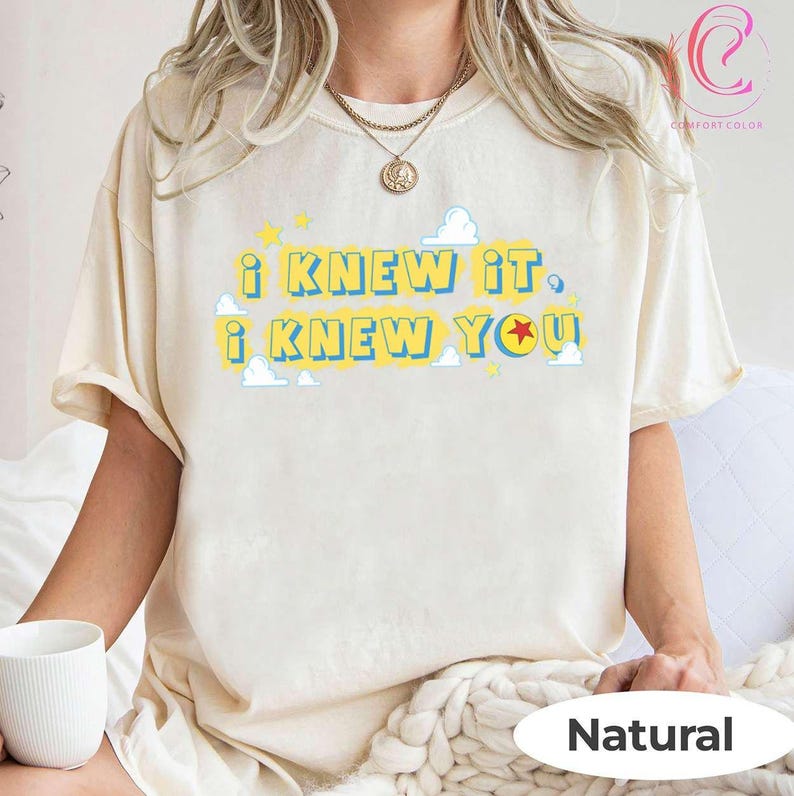 May include: A natural-colored t-shirt featuring the text "I KNEW IT, I KNEW YOU" in blue letters with yellow outlines. The design includes small yellow stars and white clouds, with a red star replacing the letter O in the word YOU.