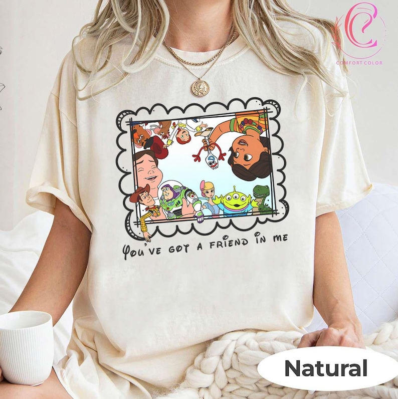 K&ouml;nnte beinhalten: Cremefarbenes T-Shirt mit einer gerahmten Grafik von Charakteren aus dem Animationsfilm "Toy Story". Die Grafik zeigt Woody, Buzz Lightyear und andere Charaktere. Der Text "You've got a friend in me" ist unter dem Rahmen aufgedruckt. Das Shirt ist mit "Natural" gekennzeichnet.