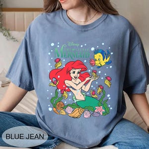 Puede incluir: Una camiseta azul con un gr&aacute;fico de Ariel de La Sirenita de Disney. Ariel tiene el pelo rojo y una cola verde, rodeada de criaturas marinas. El texto "Disney The Little Mermaid" est&aacute; encima del gr&aacute;fico. La camiseta tiene el texto "BLUE JEAN" en la parte inferior.