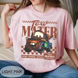 Puede incluir: Camiseta rosa claro con un dise&ntilde;o de Mate de la pel&iacute;cula Cars. El dise&ntilde;o incluye el texto "Tow Mater" y "Anytime - Anywhere - Anyplace". La camiseta tambi&eacute;n tiene el texto "Tow Mater's Towing & Salvage".