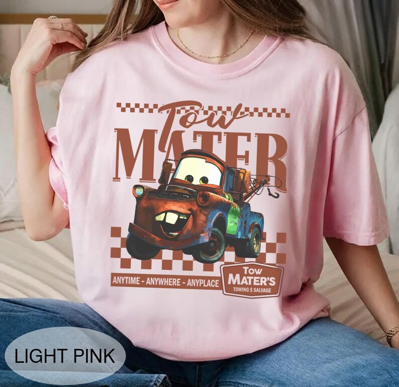 Puede incluir: Camiseta rosa claro con un dise&ntilde;o de Mate de la pel&iacute;cula Cars. El dise&ntilde;o incluye el texto "Tow Mater" y "Anytime - Anywhere - Anyplace". La camiseta tambi&eacute;n tiene el texto "Tow Mater's Towing & Salvage".