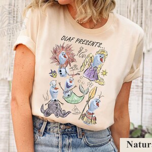 Puede incluir: Camiseta beige con una colorida ilustraci&oacute;n de Olaf de Frozen con varios disfraces, con el texto "OLAF PRESENTS...". El dise&ntilde;o incluye a Olaf como sirena, chef y otros personajes. La camiseta est&aacute; hecha de un material suave.