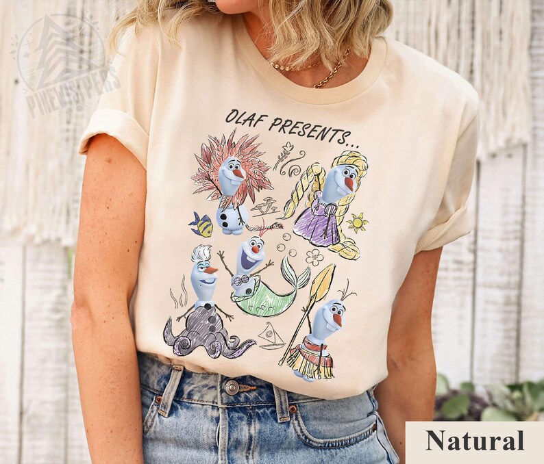 Puede incluir: Camiseta beige con una colorida ilustraci&oacute;n de Olaf de Frozen con varios disfraces, con el texto "OLAF PRESENTS...". El dise&ntilde;o incluye a Olaf como sirena, chef y otros personajes. La camiseta est&aacute; hecha de un material suave.
