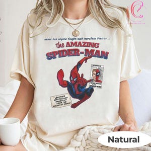 Op de afbeelding: Cr&egrave;mekleurig T-shirt met een stripboekafbeelding van Spider-Man in rood en blauw. Het shirt heeft de tekst "the AMAZING SPIDER-MAN" en andere stripboekelementen. Het shirt is onderaan voorzien van het label "Natural".