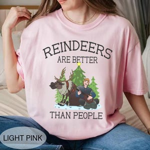 May include: Light pink t-shirt with the text "REINDEERS ARE BETTER THAN PEOPLE." The graphic features a reindeer, a man playing a stringed instrument, and Christmas trees. The shirt is a light pink color.
