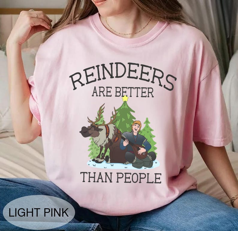May include: Light pink t-shirt with the text "REINDEERS ARE BETTER THAN PEOPLE." The graphic features a reindeer, a man playing a stringed instrument, and Christmas trees. The shirt is a light pink color.