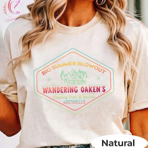 May include: A beige t-shirt with a colorful graphic that reads "BIG SUMMER BLOWOUT" above "WANDERING OAKEN'S Trading Post & Sauna ARENDELLE". The graphic features a cabin, trees, and stars. The word "Natural" is printed at the bottom.