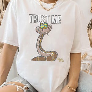 May include: White t-shirt with a cartoon image of Kaa the snake from Disney's The Jungle Book. The snake is green and brown with blue eyes and a mischievous smile. The text "TRUST ME" is printed above the snake.