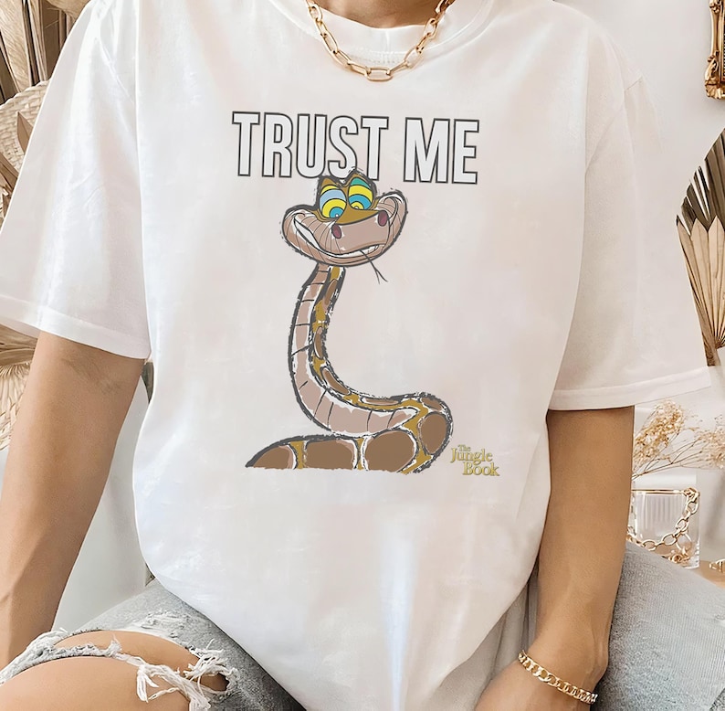 May include: White t-shirt with a cartoon image of Kaa the snake from Disney's The Jungle Book. The snake is green and brown with blue eyes and a mischievous smile. The text "TRUST ME" is printed above the snake.