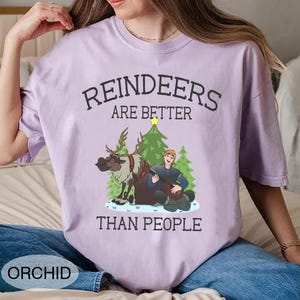 May include: Orchid-colored t-shirt with the text "REINDEERS ARE BETTER THAN PEOPLE." The graphic features a reindeer, a man playing a lute, and Christmas trees. The shirt is a comfortable, casual style.