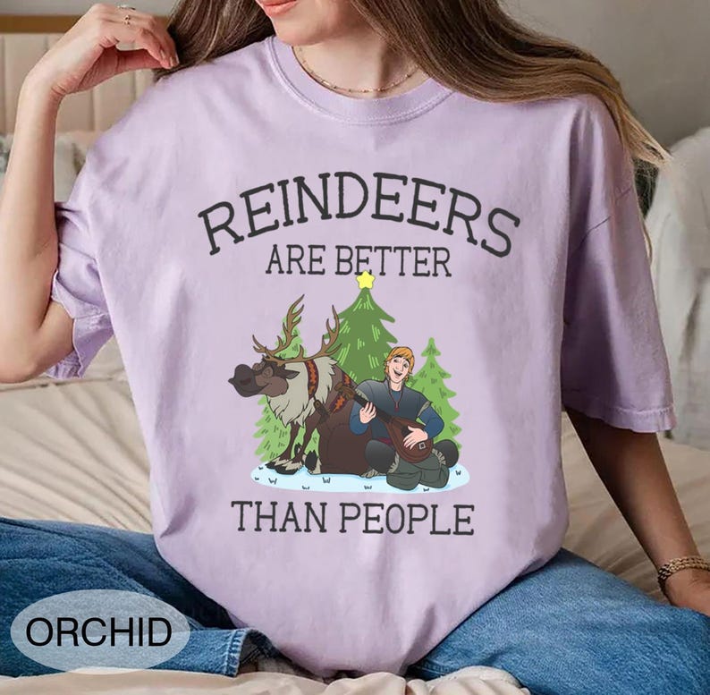 May include: Orchid-colored t-shirt with the text "REINDEERS ARE BETTER THAN PEOPLE." The graphic features a reindeer, a man playing a lute, and Christmas trees. The shirt is a comfortable, casual style.