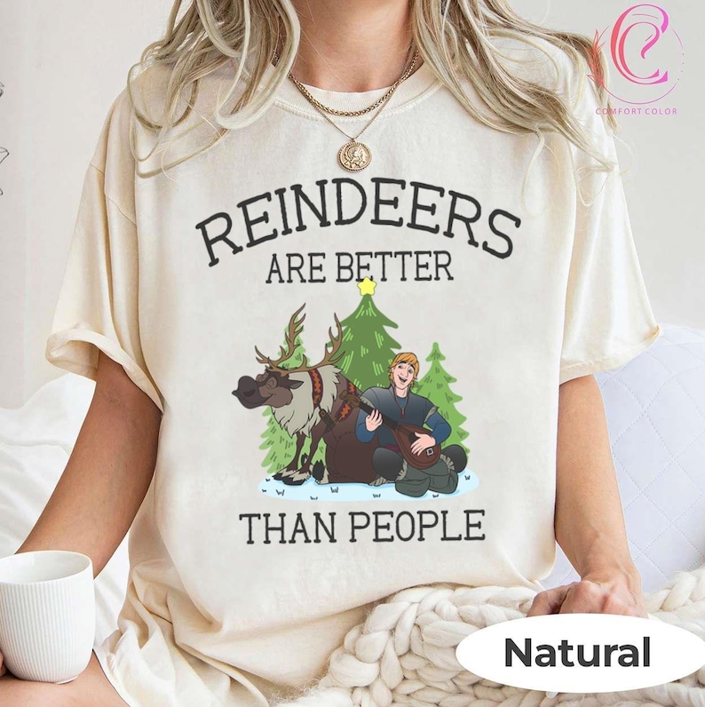 May include: Cream-colored t-shirt with the text "REINDEERS ARE BETTER THAN PEOPLE." The graphic features a cartoon reindeer, a man playing a lute, and Christmas trees. The shirt is labeled "Natural" at the bottom.