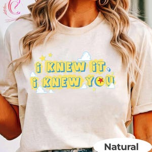 May include: A natural-colored t-shirt featuring the text "I KNEW IT I KNEW YOU" in blue letters on a yellow background. The design includes small white cloud graphics and yellow stars, with a red star replacing the letter O in the word YOU.