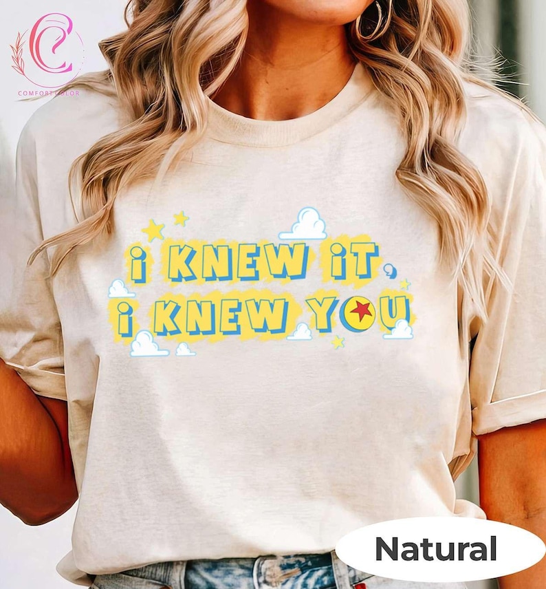 May include: A natural-colored t-shirt featuring the text "I KNEW IT I KNEW YOU" in blue letters on a yellow background. The design includes small white cloud graphics and yellow stars, with a red star replacing the letter O in the word YOU.
