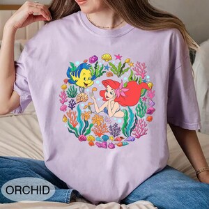 Pu&ograve; includere: T-shirt lilla chiaro con un'illustrazione colorata di Ariel e Flounder, circondati da coralli e conchiglie. La parola "ORCHID" &egrave; stampata in basso. La maglietta ha maniche corte e girocollo. Ispirata a La Sirenetta.