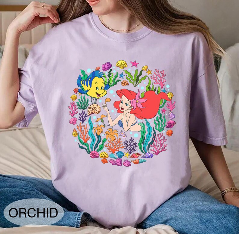 Pu&ograve; includere: T-shirt lilla chiaro con un'illustrazione colorata di Ariel e Flounder, circondati da coralli e conchiglie. La parola "ORCHID" &egrave; stampata in basso. La maglietta ha maniche corte e girocollo. Ispirata a La Sirenetta.