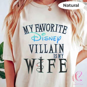 Peut inclure: T-shirt cr&egrave;me avec l'inscription "MY FAVORITE Disney VILLAIN IS MY WIFE" en noir et bleu sarcelle. Le mot "Disney" est &eacute;crit en script. Le t-shirt a une coupe d&eacute;contract&eacute;e et des manches courtes.