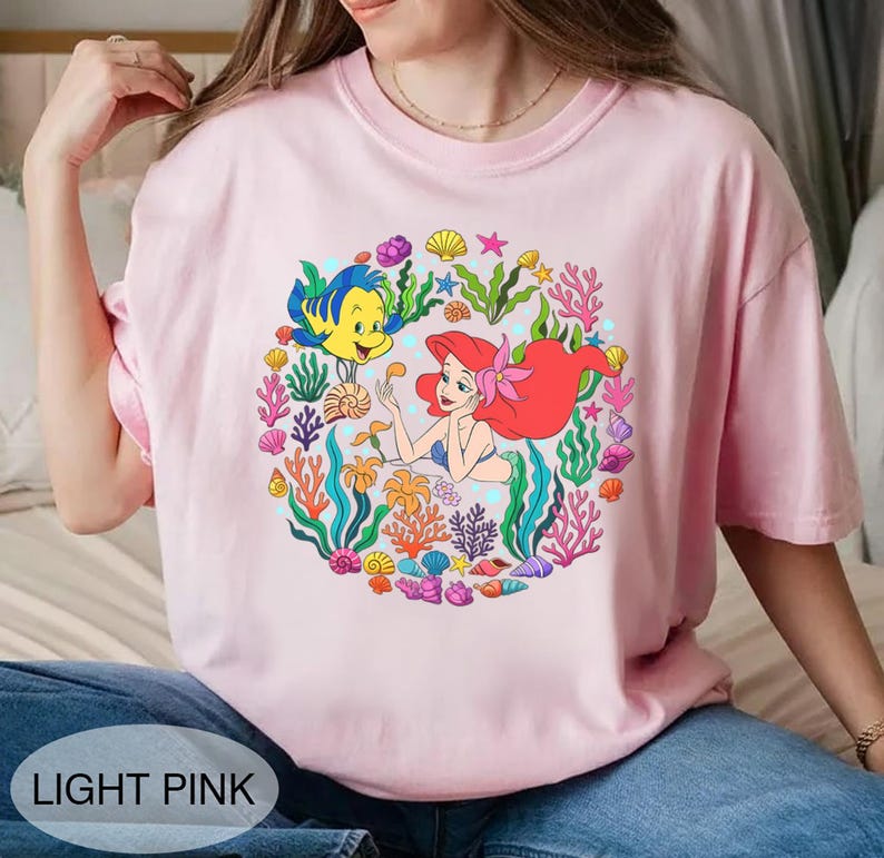 Pu&ograve; includere: T-shirt rosa chiaro con una grafica colorata di Ariel, Flounder ed elementi marini. Il design &egrave; una disposizione circolare di coralli, conchiglie e alghe. Il testo "LIGHT PINK" &egrave; in basso sulla maglietta.
