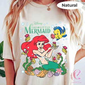 Puede incluir: Camiseta de color crema con un gr&aacute;fico colorido de Ariel de La Sirenita de Disney. Ariel, con pelo rojo y cola verde, est&aacute; rodeada de criaturas marinas. La camiseta incluye el texto "The Little Mermaid".