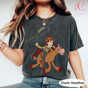May include: Dark heather gray t-shirt featuring a cartoon illustration of Woody from Toy Story riding a horse. The shirt has the name "John" in cursive script and the words "Comfort Color" and "Dark Heather" printed on it.