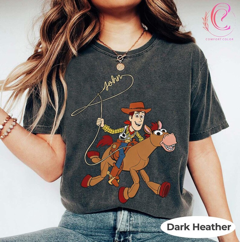 May include: Dark heather gray t-shirt featuring a cartoon illustration of Woody from Toy Story riding a horse. The shirt has the name "John" in cursive script and the words "Comfort Color" and "Dark Heather" printed on it.