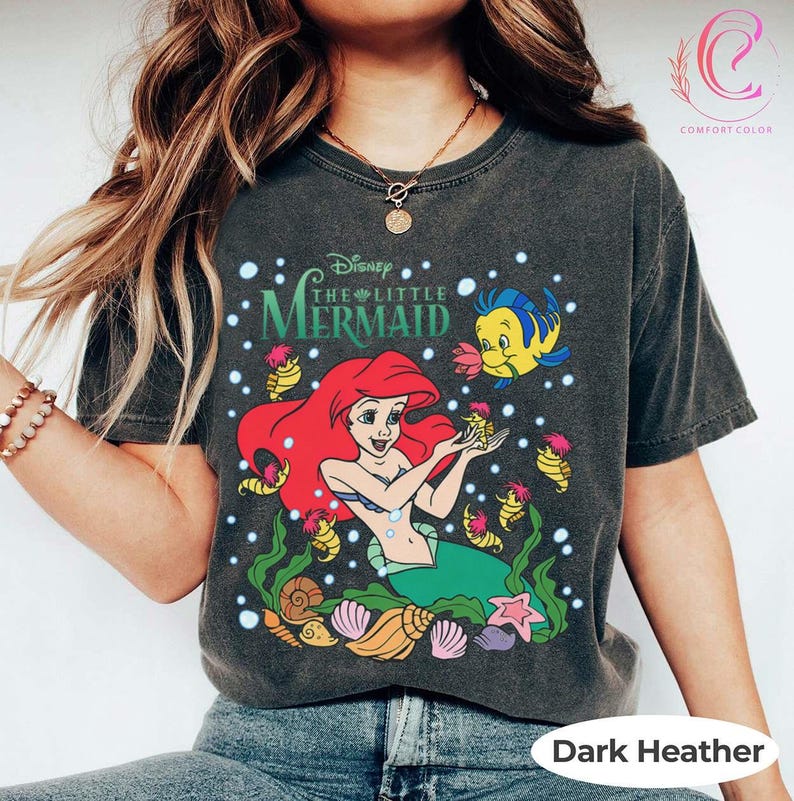 Puede incluir: Camiseta de color gris oscuro con un dise&ntilde;o de Ariel de La Sirenita de Disney. El dise&ntilde;o incluye a Ariel con pelo rojo, una cola verde y Flounder. La camiseta tambi&eacute;n tiene el texto "The Little Mermaid".