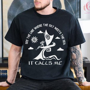 以下が含まれることがあります： 白いグラフィックデザインが施された黒いTシャツ。帆船、太陽、雲が描かれています。テキストは「SEE THE LINE WHERE THE SKY MEETS THE SEA」と「IT CALLS ME」です。