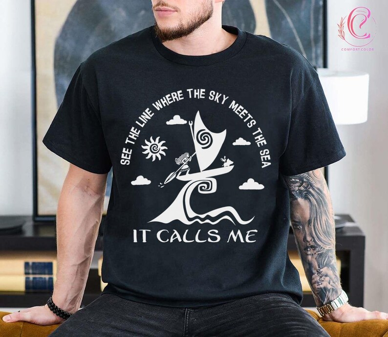 以下が含まれることがあります： 白いグラフィックデザインが施された黒いTシャツ。帆船、太陽、雲が描かれています。テキストは「SEE THE LINE WHERE THE SKY MEETS THE SEA」と「IT CALLS ME」です。