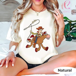May include: A cream-colored t-shirt featuring a cartoon illustration of Woody from Toy Story riding a horse. The design includes the name "John" above the cowboy and horse. The shirt is labeled "Natural" at the bottom.