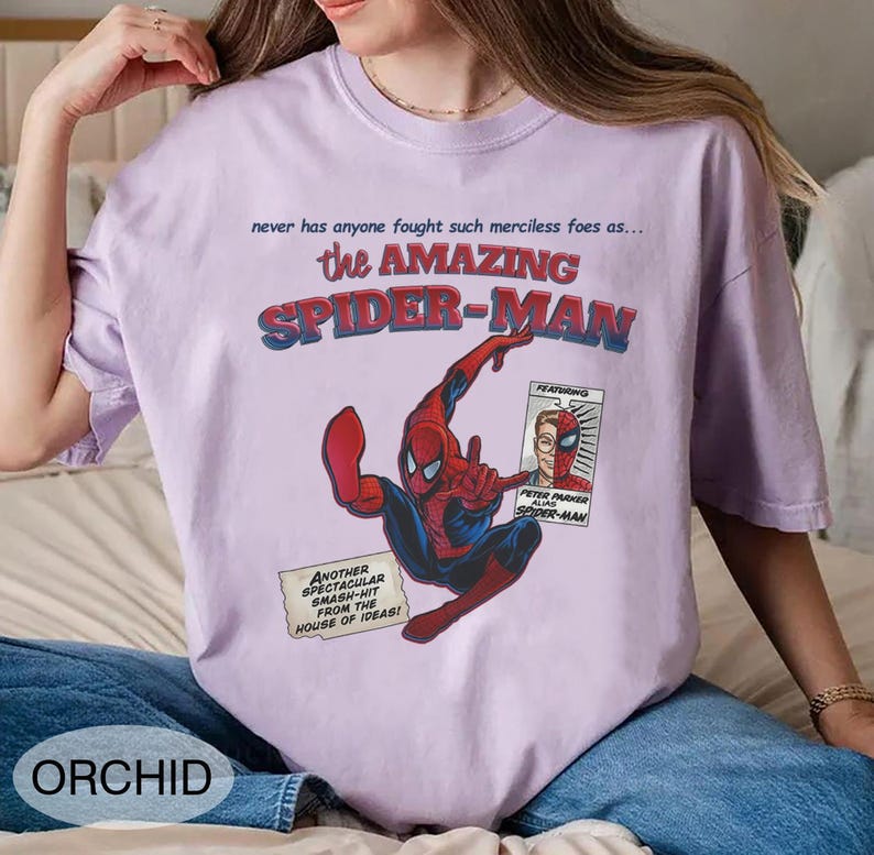 Op de afbeelding: Een lichtpaars T-shirt met een stripboekafbeelding van Spider-Man. Het ontwerp bevat de tekst "the AMAZING SPIDER-MAN" en andere stripboekelementen. Het shirt is een casual kledingstuk.