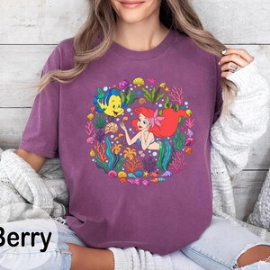 Pu&ograve; includere: T-shirt color bacca con una grafica colorata di una sirena con i capelli rossi e un pesce giallo. Il disegno &egrave; circondato da piante marine e conchiglie. La parola "Berry" &egrave; stampata in nero in basso.