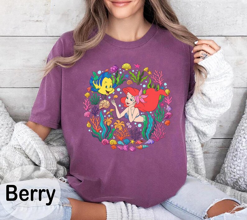 Pu&ograve; includere: T-shirt color bacca con una grafica colorata di una sirena con i capelli rossi e un pesce giallo. Il disegno &egrave; circondato da piante marine e conchiglie. La parola "Berry" &egrave; stampata in nero in basso.