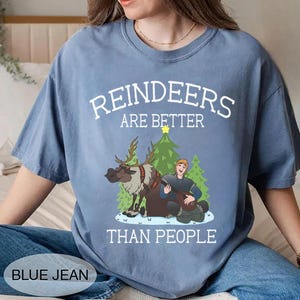 May include: A blue jean-colored t-shirt with the text "REINDEERS ARE BETTER THAN PEOPLE" in white. The graphic features a cartoon reindeer, a man playing a lute, and Christmas trees. The shirt is a casual, unisex style.
