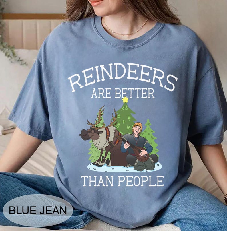 May include: A blue jean-colored t-shirt with the text "REINDEERS ARE BETTER THAN PEOPLE" in white. The graphic features a cartoon reindeer, a man playing a lute, and Christmas trees. The shirt is a casual, unisex style.