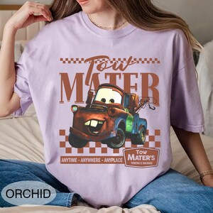 Puede incluir: Camiseta color orqu&iacute;dea con una imagen de dibujos animados de Tow Mater de la pel&iacute;cula Cars. El dise&ntilde;o incluye el texto "Tow Mater" en marr&oacute;n, un patr&oacute;n de cuadros y la frase "ANYTIME - ANYWHERE - ANYPLACE".