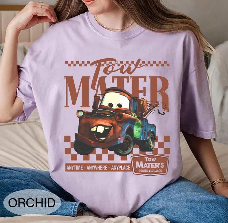 Puede incluir: Camiseta color orqu&iacute;dea con una imagen de dibujos animados de Tow Mater de la pel&iacute;cula Cars. El dise&ntilde;o incluye el texto "Tow Mater" en marr&oacute;n, un patr&oacute;n de cuadros y la frase "ANYTIME - ANYWHERE - ANYPLACE".