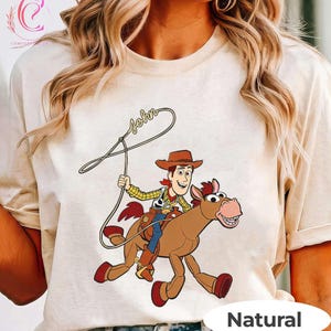 May include: A natural-colored t-shirt featuring a cartoon illustration of a cowboy riding a horse, holding a lasso. The cowboy wears a hat, a yellow plaid shirt, and blue pants. The word "John" is written above the lasso.