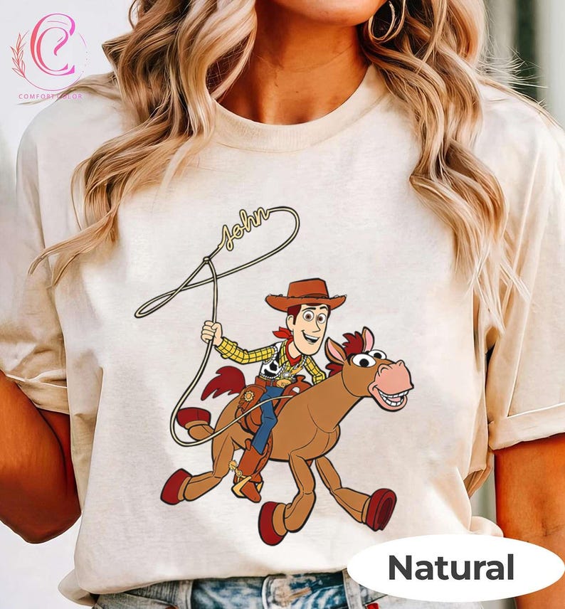 May include: A natural-colored t-shirt featuring a cartoon illustration of a cowboy riding a horse, holding a lasso. The cowboy wears a hat, a yellow plaid shirt, and blue pants. The word "John" is written above the lasso.