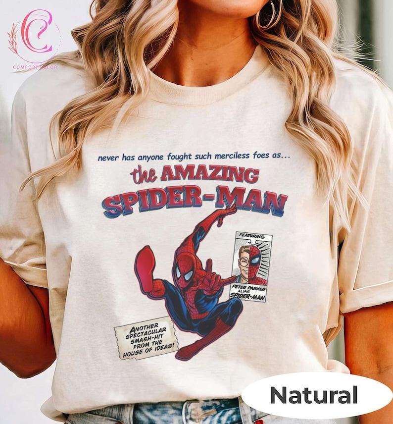 Op de afbeelding: Beige T-shirt met een stripboekafbeelding van Spider-Man in rood en blauw. De tekst "the AMAZING SPIDER-MAN" is in rood en blauw. Extra tekst is onder meer "never has anyone fought such merciless foes as..." en "ANOTHER SPECTACULAR SMASH-HIT FROM THE HOUSE OF IDEAS!"