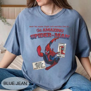Op de afbeelding: Lichtblauw T-shirt met een Spider-Man-afbeelding. Het ontwerp bevat de tekst "the AMAZING SPIDER-MAN" en "PETER PARKER alias SPIDER-MAN". Het shirt heeft ook de tekst "BLUE JEAN".
