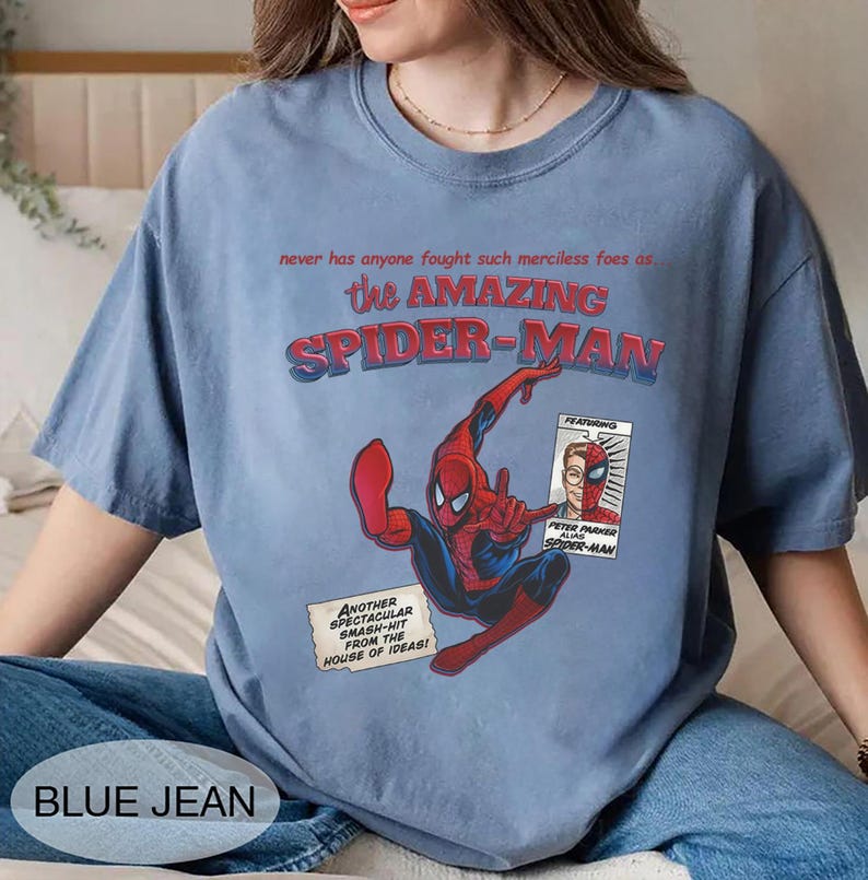 Op de afbeelding: Lichtblauw T-shirt met een Spider-Man-afbeelding. Het ontwerp bevat de tekst "the AMAZING SPIDER-MAN" en "PETER PARKER alias SPIDER-MAN". Het shirt heeft ook de tekst "BLUE JEAN".