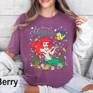 Puede incluir: Camiseta de manga larga color bayas con un gr&aacute;fico de Ariel de La Sirenita de Disney. El dise&ntilde;o incluye a Ariel, Flounder y varios elementos marinos. Tambi&eacute;n es visible el texto "Disney The Little Mermaid".