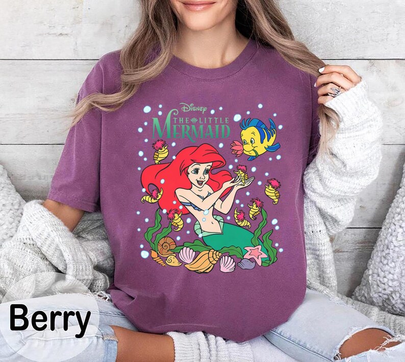 Puede incluir: Camiseta de manga larga color bayas con un gr&aacute;fico de Ariel de La Sirenita de Disney. El dise&ntilde;o incluye a Ariel, Flounder y varios elementos marinos. Tambi&eacute;n es visible el texto "Disney The Little Mermaid".