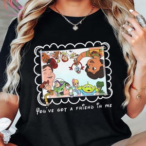 K&ouml;nnte beinhalten: Schwarzes T-Shirt mit einem farbenfrohen Grafikdesign von "Toy Story"-Charakteren in einem wei&szlig;en, wellenf&ouml;rmigen Rahmen. Der Text "You've got a friend in me" ist darunter in Wei&szlig; gedruckt. Ein lustiges, l&auml;ssiges Oberteil.