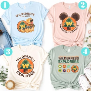 Peut inclure: Quatre t-shirts à manches courtes en bleu clair, rose clair, crème et vert sauge. Chaque t-shirt présente un motif Wilderness Explorers avec un arbre, les lettres WE et divers badges. Le t-shirt bleu clair inclut un chien.