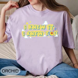 May include: A light purple short-sleeve t-shirt featuring the yellow text I KNEW IT I KNEW YOU with blue outlines. The design includes small white clouds and yellow stars, with a red and yellow ball replacing the letter O in YOU.