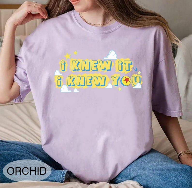 May include: A light purple short-sleeve t-shirt featuring the yellow text I KNEW IT I KNEW YOU with blue outlines. The design includes small white clouds and yellow stars, with a red and yellow ball replacing the letter O in YOU.