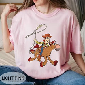 May include: Light pink t-shirt featuring a cartoon cowboy riding a brown horse with a rope. The cowboy wears a brown hat, yellow shirt, and blue pants. The word "John" is written above the cowboy. The shirt has the words "LIGHT PINK" at the bottom.