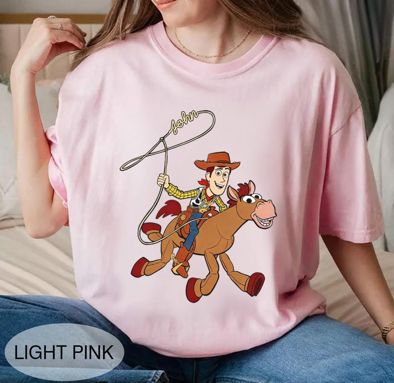 May include: Light pink t-shirt featuring a cartoon cowboy riding a brown horse with a rope. The cowboy wears a brown hat, yellow shirt, and blue pants. The word "John" is written above the cowboy. The shirt has the words "LIGHT PINK" at the bottom.