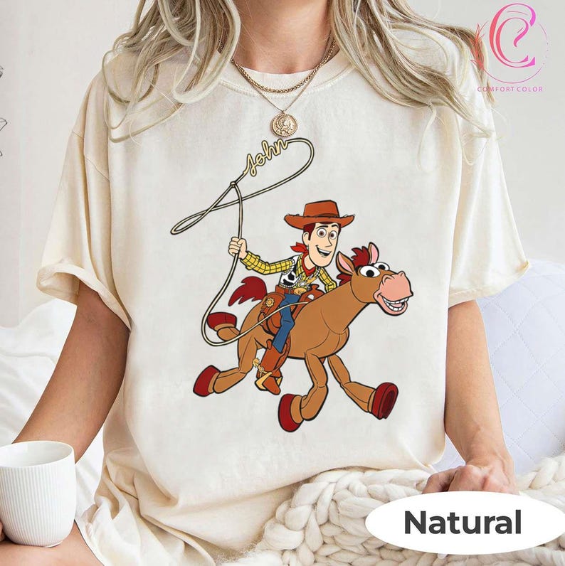 May include: Cream-colored t-shirt featuring a cartoon illustration of Woody from Toy Story riding a horse. The design includes the name "John" in script and the word "Natural" at the bottom. The shirt is a Comfort Colors brand.